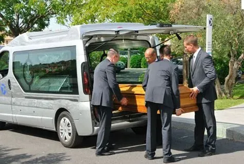 Quatre porteurs chargent avec respect un cercueil en bois dans le corbillard.